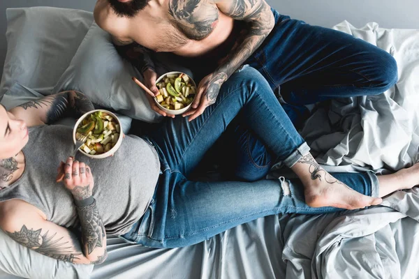 Cropped image of pregnant tattooed girlfriend eating salad with boyfriend on bed — Stock Photo