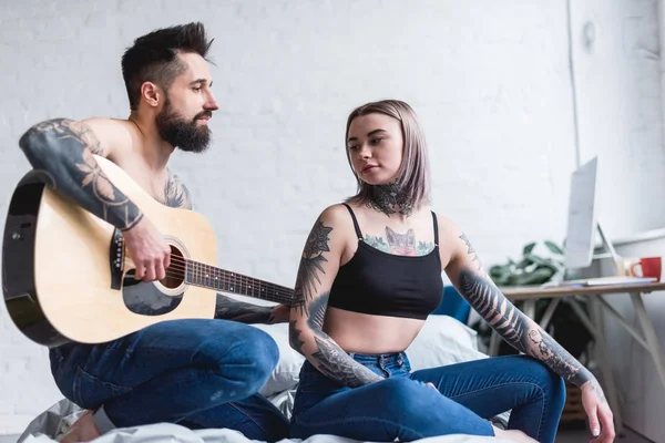Tattooed boyfriend playing guitar for girlfriend on bed — Stock Photo