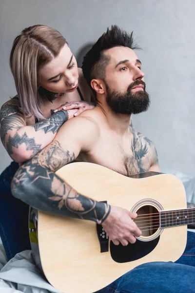 Novio tocando la guitarra para la novia tatuada en casa - foto de stock