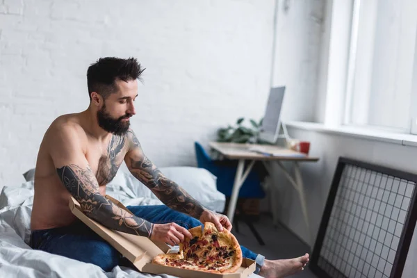 Hemdlos gutaussehender tätowierter Mann isst Pizza im Schlafzimmer — Stockfoto