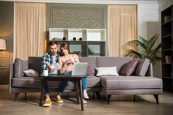 Pareja libro de lectura en la sala de estar con ordenador portátil en la mesa en la acogedora sala de estar - foto de stock