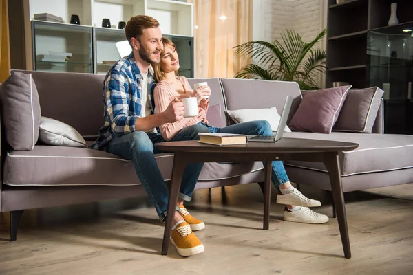 Vista lateral de hombre con novia sentado en el sofá con tazas y viendo el ordenador portátil en la sala de estar moderna - foto de stock