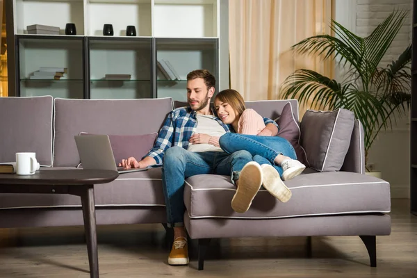 Mujer abrazando novio mientras que el uso de la computadora portátil en la sala de estar con diseño moderno - foto de stock