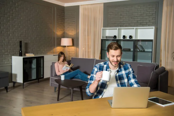 Hombre con la taza en la mano usando el ordenador portátil, mientras que la mujer que lee el libro en el sofá en la sala de estar en diseño moderno - foto de stock