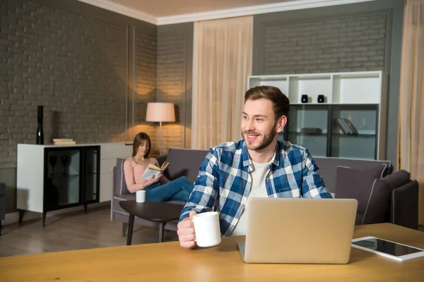 Uomo sorridente a tavola con computer portatile e donna lettura libro sul divano in soggiorno moderno — Foto stock