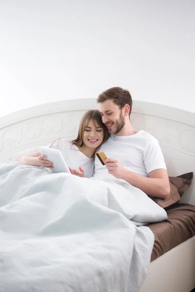 Mujer sonriente usando tableta digital con su novio sosteniendo la tarjeta de crédito en el dormitorio acogedor - foto de stock