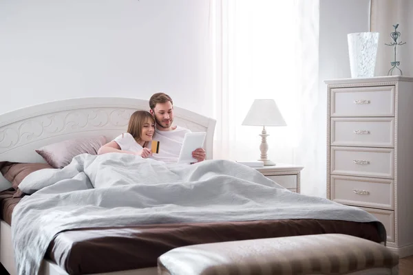 Jeune couple couché au lit et faisant du shopping en ligne dans la chambre moderne — Photo de stock