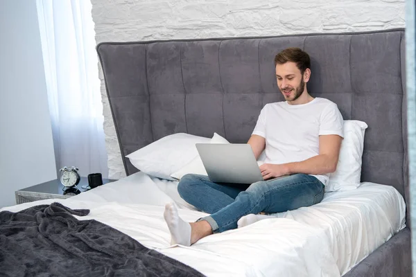 Maschio sorridente utilizzando il computer portatile in camera da letto con interni moderni — Foto stock