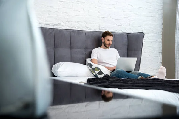 Vista de nível de superfície do jovem sentado na cama com laptop no quarto moderno — Fotografia de Stock