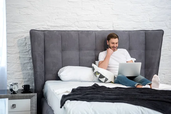 Hombre joven feliz usando el ordenador portátil en el dormitorio moderno acogedor - foto de stock