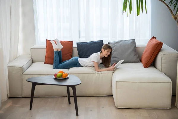 Vista laterale della giovane donna sdraiata sul divano e utilizzando tablet digitale in soggiorno con interni moderni — Foto stock