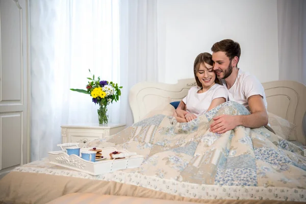 Casal feliz sentado na cama com café da manhã na bandeja no quarto acolhedor — Fotografia de Stock