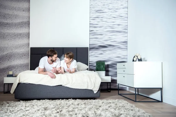 Pareja sonriente hablando y acostada en la cama en un acogedor dormitorio con interior moderno - foto de stock