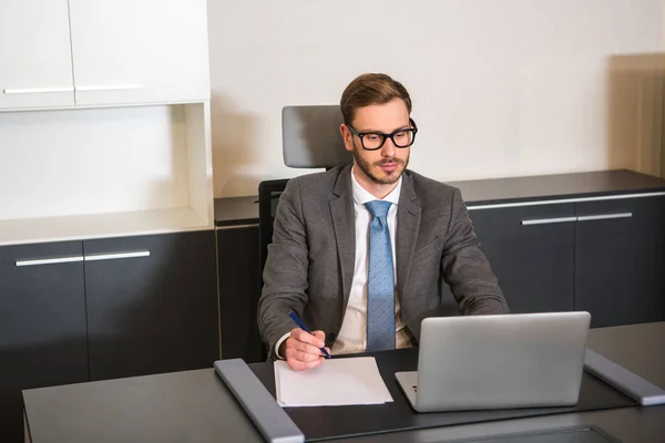 Vista frontal del hombre de negocios sentado en la mesa con el ordenador portátil y la pluma en la mano - foto de stock