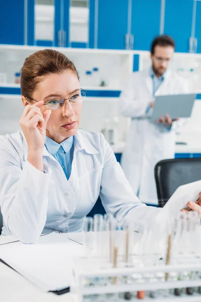 Foyer sélectif de la femme scientifique en utilisant la tablette sur le lieu de travail avec collègue derrière en laboratoire — Photo de stock