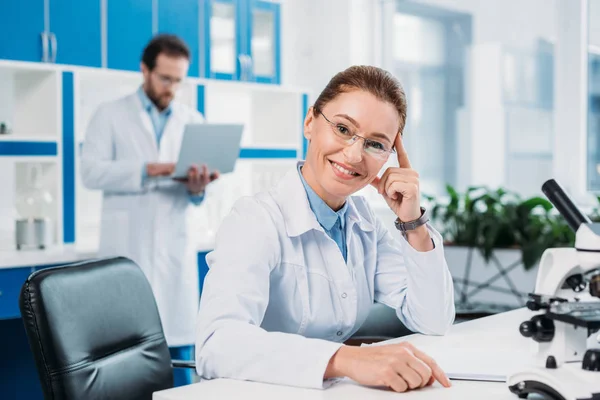 Selektiver Fokus einer lächelnden Wissenschaftlerin mit Brille am Arbeitsplatz mit einem Kollegen im Labor dahinter — Stockfoto