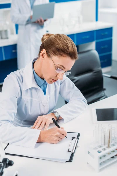 Enfoque selectivo de la científica femenina haciendo notas en bloc de notas con su colega detrás en el laboratorio - foto de stock