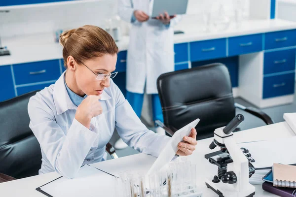 Foyer sélectif de la femme scientifique en utilisant la tablette sur le lieu de travail avec collègue derrière en laboratoire — Photo de stock