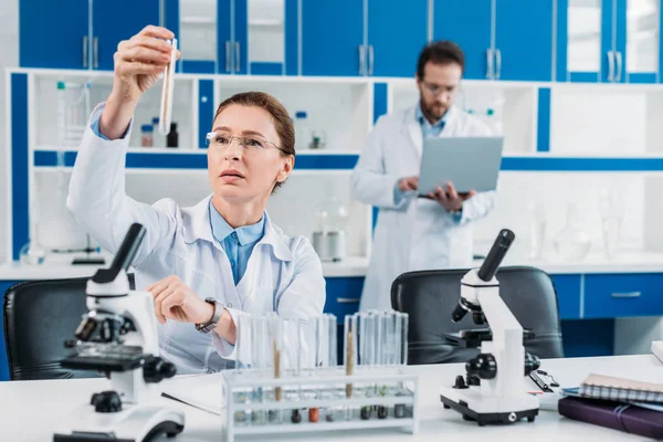 Enfoque selectivo de la mujer científica mirando tubo con reactivo en la mano con colega detrás en el laboratorio - foto de stock