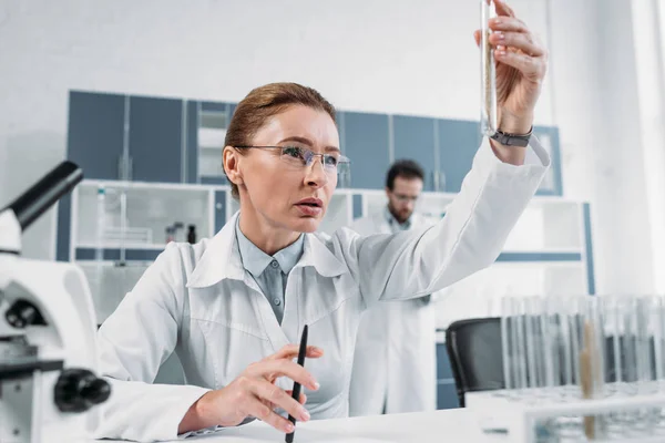 Вибірковий фокус жінки-вченого, дивлячись на трубку з реагентом в руці з колегою позаду в лабораторії — стокове фото