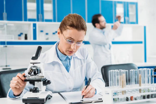 Selektive Fokussierung der Wissenschaftlerin, die am Arbeitsplatz Notizen im Notizblock macht, während die Kollegin im Labor dahinter steht — Stockfoto