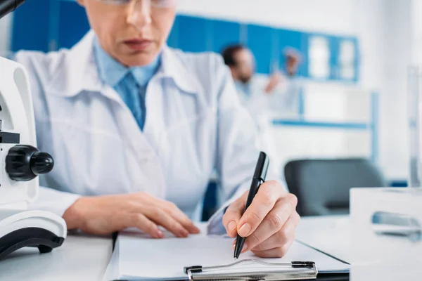 Partial view of female scientist making notes in notepad at workplace with colleague behind in lab — Stock Photo