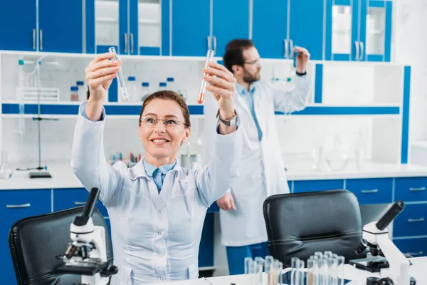 Messa a fuoco selettiva di ricercatrice donna guardando tubi con reagenti in mano con collega dietro in laboratorio — Foto stock