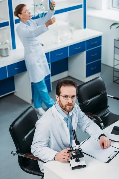 Vista de alto ángulo del científico en bata blanca en el lugar de trabajo con microscopio en laboratorio - foto de stock