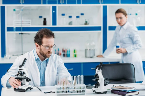 Foco seletivo do cientista em casaco branco e óculos no local de trabalho com microscópio e colega atrás em laboratório — Fotografia de Stock