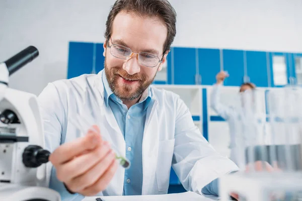 Foco seletivo do cientista sorridente em casaco branco e óculos olhando para o frasco com reagente na mão em laboratório — Fotografia de Stock