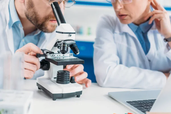 Visão parcial de pesquisadores científicos em casacos brancos trabalhando juntos no local de trabalho com microscópio em laboratório — Fotografia de Stock
