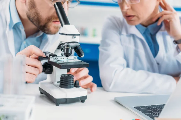 Visión parcial de los investigadores científicos en batas blancas que trabajan juntos en el lugar de trabajo con microscopio en laboratorio - foto de stock