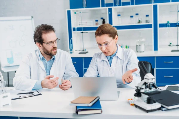 Portrait of scientists in lab coats and eyeglasses working together at workplace with laptop in lab — Stock Photo
