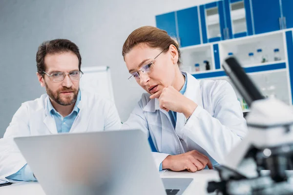 Portrait of scientists in lab coats and eyeglasses working together at workplace with laptop in lab — Stock Photo