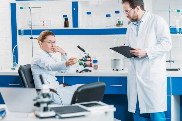 Pesquisador científico olhando para frasco com reagente na mão com colega por perto em laboratório — Fotografia de Stock