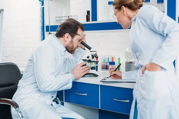 Scientific researcher looking through microscope on reagent with colleague near by in laboratory — Stock Photo