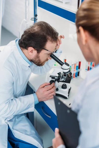 Científico en bata de laboratorio mirando a través del microscopio en reactivo con colega cerca en laboratorio - foto de stock