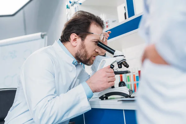 Wissenschaftler im Laborkittel schaut durch das Mikroskop auf Reagenz mit einem Kollegen in der Nähe im Labor — Stockfoto