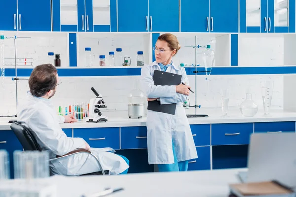Vista parcial de la científica en gafas con bloc de notas en las manos mirando a su colega en el lugar de trabajo en el laboratorio - foto de stock
