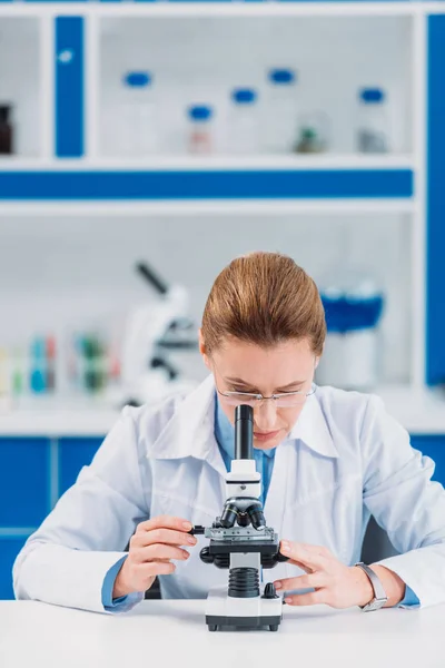 Científica femenina en anteojos que mira el reactivo a través del microscopio en laboratorio - foto de stock