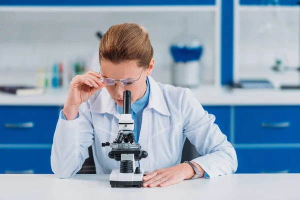 Cientista feminina em óculos procurando reagente através de microscópio em laboratório — Fotografia de Stock