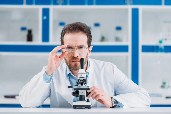 Retrato de científico en bata blanca y anteojos con microscopio en laboratorio - foto de stock