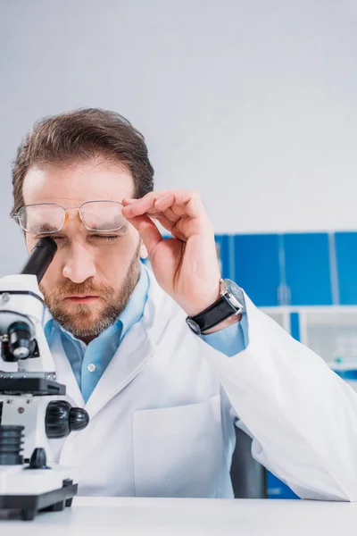 Ученый в белом халате и очках, смотрящий через микроскоп на реагент в лаборатории — стоковое фото