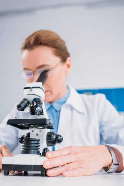 Selective focus of female scientist in lab coat and eyeglasses looking through microscope on reagent in lab — Stock Photo