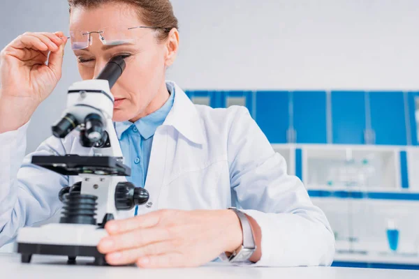 Científica en bata de laboratorio y anteojos mirando a través del microscopio en reactivo en laboratorio - foto de stock