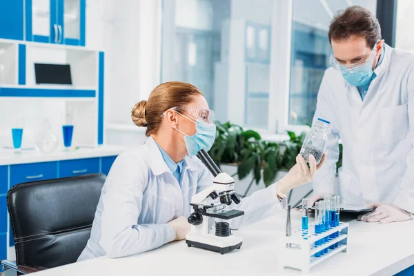 Científicos con batas blancas y gafas que trabajan con reactivos y microscopio en laboratorio - foto de stock