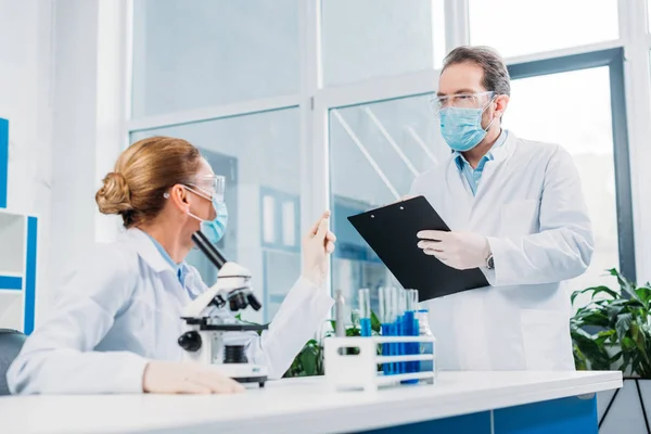 Científicos con batas blancas y gafas que trabajan con reactivos y microscopio en laboratorio - foto de stock