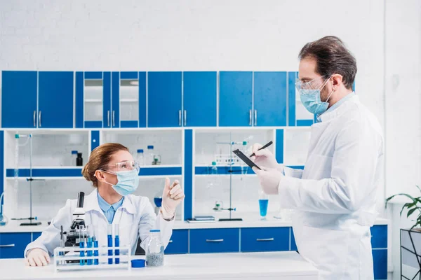 Científicos con batas blancas y gafas que trabajan con reactivos y microscopio en laboratorio - foto de stock