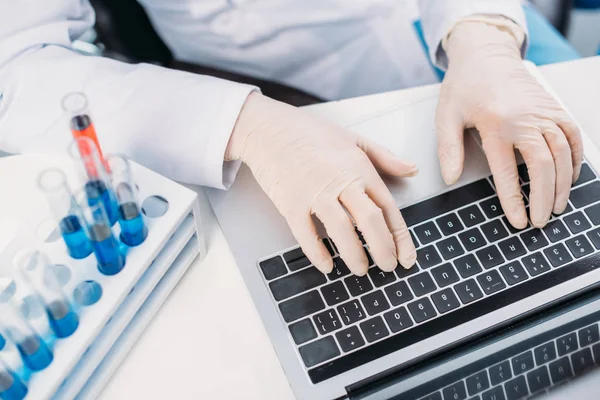 Partial view of scientist in lab coat and medical gloves typing on laptop at workplace with reagents — Stock Photo