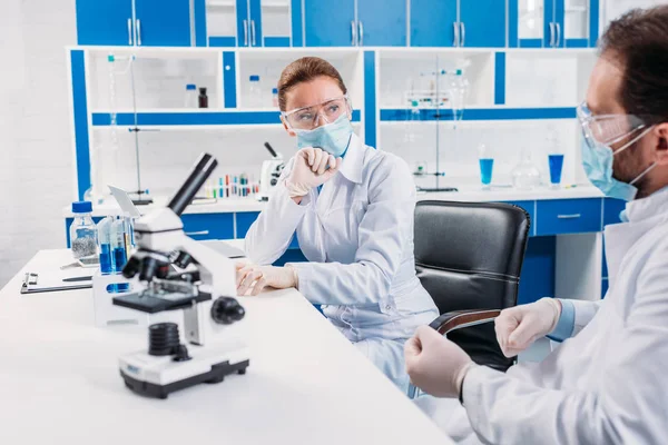 Scientific researchers in medical masks and goggles having discussion at workplace with microscope in lab — Stock Photo